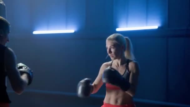 キックボクシングの訓練だ ボクシングの手袋の女性は青い光と暗いジムで彼女のコーチとボクシングです 女性キックボクサーと男性トレーナーのトレーニングスパーリング 女戦士の練習パンチとキック — ストック動画