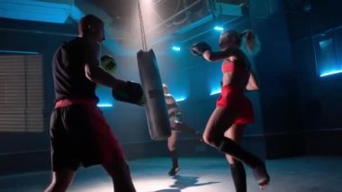 Kickboks eğitimi. Ön planda kadın boksör antrenörle yumruk ve tekme atmayı öğretiyor. Arka planda, bir bayan atlet kum torbasına vurma alıştırması yapıyor. Kickboxerlar siyah spor salonunda boks eldivenleriyle