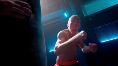 Genç erkek boksör yumruk atıyor ve bacakları tekmeliyor. Koyu renk spor salonunda mavi ışıkla kum torbasını yumrukluyor. Çıplak gövdeli bir sporcu antrenmanda yumruk pratiği yapar. Boks, kickboks, kendini savunma kavramı