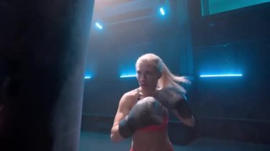 Koyu renk spor salonunda mavi ışıklı kadın boksör eğitimi. Siyah boks eldivenli bir kadın antrenörün tuttuğu kum torbasında yumruk pratiği yapıyor. Kickboks antrenmanı. Sporcu bir kadın. Ağır çekim hazır, 4K 'de.