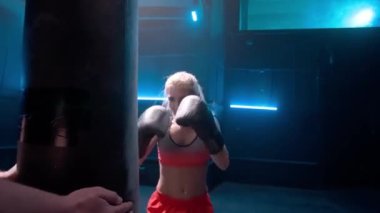 Koyu renk spor salonunda mavi ışıklı kadın boksör eğitimi. Siyah boks eldivenli bir kadın kum torbasında yumruk pratiği yapıyor. Kickboks antrenmanı. Sporcu bir kadın. Yavaş çekim hazır, 4K 59.94fps 'de..