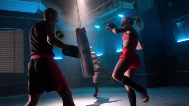 キックボクシングの訓練だ フォアグラウンドの女性戦闘機でパンチとトレーナーでキックを訓練します 背景には 女性アスリートがパンチングバッグを練習している 暗いジムでのボクシンググローブのキックボクサー — ストック動画