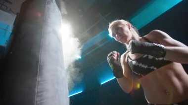 Genç bir kadının kum torbasına güçlü yumruklar atmasına düşük görüş açısı. Siyah boks bandajlı kadın sporcu elleri koyu renk spor salonunda mavi ışıkla kum torbasına saldırıyor. Boks veya