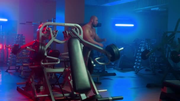 赤身裸体的健美运动员正在热身 准备在运动模拟器上进行力量训练 身穿黑色短裤的男人带着各种装备 蓝色的哑铃架在体育馆里走来走去 — 图库视频影像