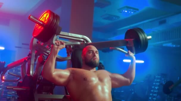 男性健美运动员赤身裸体坐在运动模拟器上 运动员的运动技术是将胳膊向上挤压 然后从运动中休息 男人正在把胸部的肌肉抽出来 — 图库视频影像