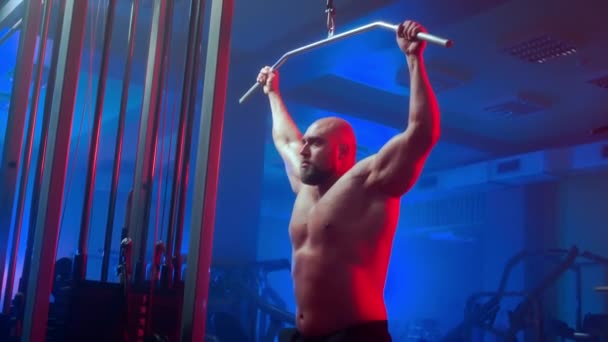 具有抽吸肌肉的健美运动员正在运动模拟器上进行上块拉运动 赤身裸体的男人在体育馆用蓝红相间的力量拉起酒吧 — 图库视频影像