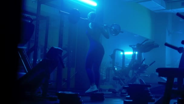 運動能力の女性の暗いシルエットのリアビューは 青の光と暗いジムでバーベルでスクワットしています スポーツ女性のボディビルダーは お尻と太ももの筋肉のための演習を行っています — ストック動画