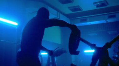 Dark silhouette of a man with a muscular body is fixing a sports athletic belt on the lower abdomen. Athlete bodybuilder is preparing for weightlifting strength training in a dark gym with blue light