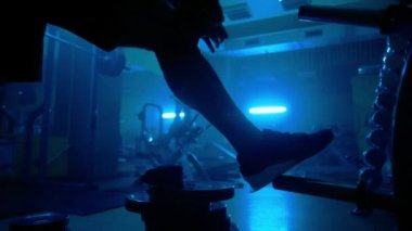Dark silhouette of a muscular man is bandaging his knees with sports bandages. Athlete bodybuilder is preparing for strength training. Strongman is posing in dark gym with blue light against