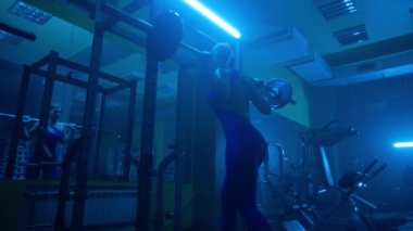 Side view of dark silhouette of woman with slender body is squatting with barbell in dark gym with blue light. Female bodybuilder is doing exercises for buttocks muscles and looking in mirror to