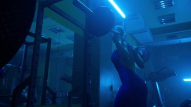 Side view of dark silhouette of woman with slender body is squatting with barbell in dark gym with blue light. Female bodybuilder is doing exercises for buttocks muscles and looking in mirror to