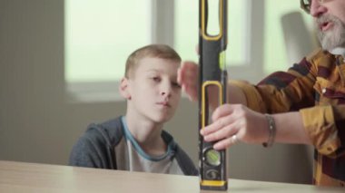 Büyükbaba torununa su seviyesinin nasıl işlediğini anlatıyor ve öğretiyor. Yaşlı bir adam ve genç bir adam pencerenin arka planında bir dairede oturuyorlar ve başparmaklarını göstermekten memnunlar.