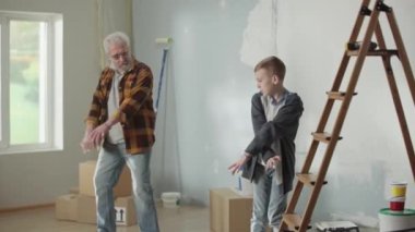 Grandson is teaching grandpa how to dance and showing modern dance moves with his hands. Elderly man and young guy are dancing merrily and smiling joyfully against the backdrop of an apartment being