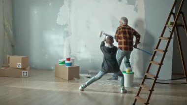 Grandson taking photo with mobile phone and filming a video of grandpa painting a wall white with a paint roller. Elderly man doing repairs in apartment and posing for photo for memory. Concept of