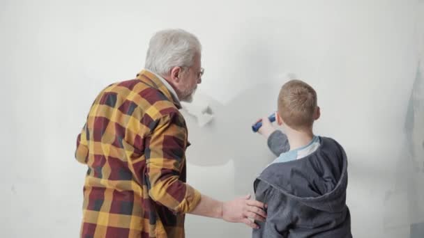 从爷爷和孙子的后面看 是用油漆滚筒把墙漆成白色的 一位老人正在教一个年轻人如何修理和提供建议 维修的概念 — 图库视频影像