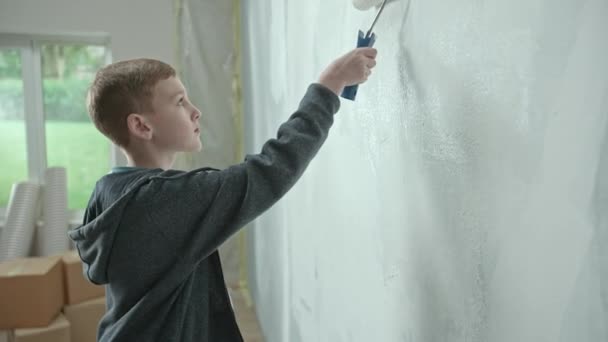 身穿蓝色连帽衫的小男孩正在用油漆辊把墙壁涂成白色 青少年检查墙壁 勤勤恳恳地沿着墙壁跑来跑去 公寓维修 学习的概念 — 图库视频影像