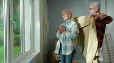 An elderly man wraps plaid around an older woman with cup of tea in his hands. Aged couple of pensioners dreaming about something, standing at the window in a cozy home environment