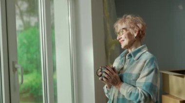 An elderly man wraps plaid around an older woman with cup of tea in his hands. Aged couple of pensioners dreaming about something, standing at the window in a cozy home environment