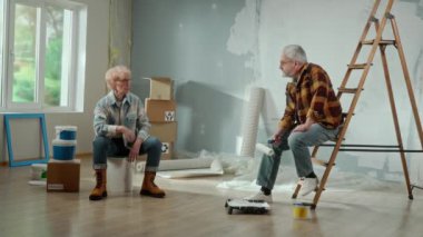 Tired elderly man is calling a master to repair an apartment using a mobile phone. Couple of aged is rejoicing at the solved problem and the help of the master. Ladder, cardboard boxes and window in