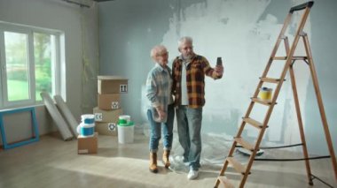 Elderly man and woman are talking on a video call using their mobile phone. Aged couple communicates with someone and showing repairs in the apartment, ladder, cardboard boxes, a window and wallpaper