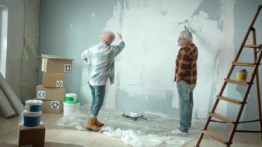 Elderly man painting wall with white paint using paint roller. Older woman leads painting process and gives valuable advice. Couple of pensioners making repairs to their apartment, against backdrop of