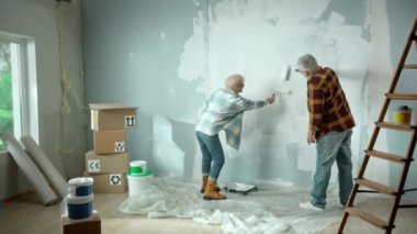 Elderly man and woman are painting wall with white paint using paint rollers. Couple of pensioners is making repairs to their apartment, in the background of window, stepladder, cardboard boxes