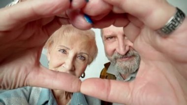 Portrait of a happy elderly couple is making hand heart, looking at camera, smiling through finger frame during zooming camera. Older man and woman are showing sign of heart, love, tenderness