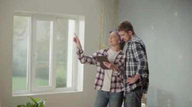 Happy young couple in checkered shirts using tablet to plan renovations in their apartment. Man and woman communicate, discuss a project for repairing room and point finger in different directions