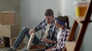 Young couple sits on oilcloth on floor and enjoys eating pizza while relaxing. Man and woman in checkered shirts and jeans are having fast food lunch against backdrop of bright room during renovation