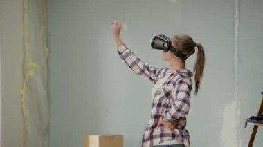 Young woman in virtual reality headset furnishes interior of room. Female in checkered shirt and jeans makes choice of wallpaper, furniture, curtains using virtual technology. Concept of modern