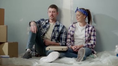 Young couple sits on oilcloth on floor and enjoys eating pizza while relaxing. Man and woman in checkered shirts and jeans are having fast food lunch against backdrop of bright room during renovation