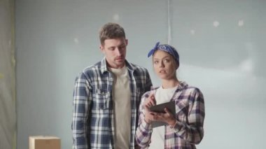Happy young couple in checkered shirts using tablet to plan renovations in their apartment. Man and woman communicate, discuss a project for repairing room and point finger in different directions