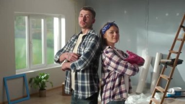 Portrait of woman in pink rubber gloves and man crossing their arms, looking seriously at camera and smiling. Young couple in plaid shirts posing against backdrop of room in process of being renovated