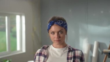 Portrait of woman in pink rubber gloves crossing her arms, looking seriously at camera and smiling. Young female in checkered shirt and blue bandana posing against backdrop of room in process of being