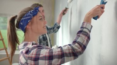 Young couple are painting wall with white paint using paint rollers against backdrop of window with bright sunlight. Pretty woman and man in checkered shirts enjoying renovations in apartment and