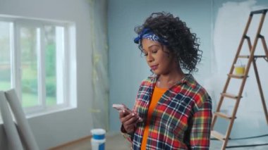 Young African American woman is looking at phone screen and smiling. Portrait of black female looking through information in smartphone against the background of an apartment in process of renovation