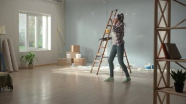 Büyük kablosuz beyaz kulaklıklı, müzikten ve danstan hoşlanan genç bir kadın. Kareli gömlekli ve kotlu bir kadın mikrofona sarılmış gibi neşeli bir şekilde boya silindirine şarkı söylüyor. Neşeli ruh hali kavramı