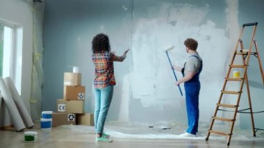 African American woman is giving directions to male house painter painting wall with white paint using long paint roller. Repairman in blue construction overalls doing repairs in an apartment. Concept