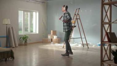 Young man in big wireless white headphones enjoying music and dancing. Male in checkered shirt and jeans cheerfully singing into paint roller as if into microphone. Concept of cheerful mood during