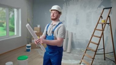 Male foreman is looking through large sheet with an apartment plan and thinking over repair project in flat. Redhead man with beard in blue construction overalls and white helmet is planning finishing