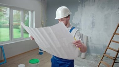 Male foreman is looking through large sheet with an apartment plan and thinking over repair project in flat. Redhead man with beard in blue construction overalls and white helmet is planning finishing