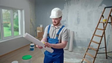 Male foreman is looking through large sheet with an apartment plan and thinking over repair project in flat. Redhead man with beard in blue construction overalls and white helmet is planning finishing