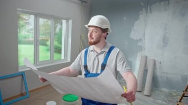 Male foreman is looking through large sheet with an apartment plan and thinking over repair project in flat. Redhead man with beard in blue construction overalls and white helmet is planning finishing