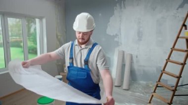 Male foreman is looking through large sheet with an apartment plan and thinking over repair project in flat. Redhead man with beard in blue construction overalls and white helmet is planning finishing