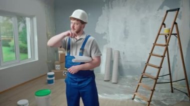 Male foreman is looking through large sheet with an apartment plan and thinking over repair project. Redhead man with beard in blue construction overalls and white helmet in anger throws away sheet