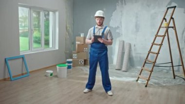 Male foreman in blue construction overalls and white helmet is planning renovation project in an apartment using tablet. Redhead master man with beard thinking, viewing and visualizing ideas against