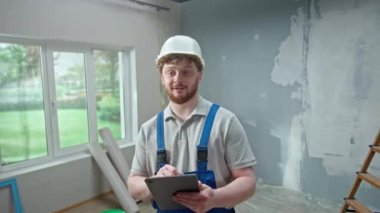 Male foreman in blue construction overalls and white helmet is planning renovation project in an apartment using tablet. Redhead master man with beard thinking, viewing and visualizing ideas against