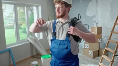 Foreman or electrician in blue overalls, white helmet and coil of black cable in hands. Redhead man is planning repairs in an apartment against backdrop of ladder, cardboard boxes and window. Concept