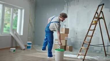 Foreman or painter in blue construction overalls and white helmet with bucket of paint in his hands. Redhead man taking paint roller and planning to paint walls in renovated apartment. Concept of