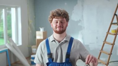 Repairman worker in blue construction overalls showing bunch of keys and smiling. Portrait of redhead man posing against backdrop of apartment in process of renovation, ladder, cardboard boxes, window